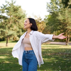 Westside Behavioral Care ADHD ADD Westside Behavioral Care ADHD and ADD Section - Young woman in jeans and a white shirt smiling with her arms outstretched in a green park surrounded by trees on a sunny day.