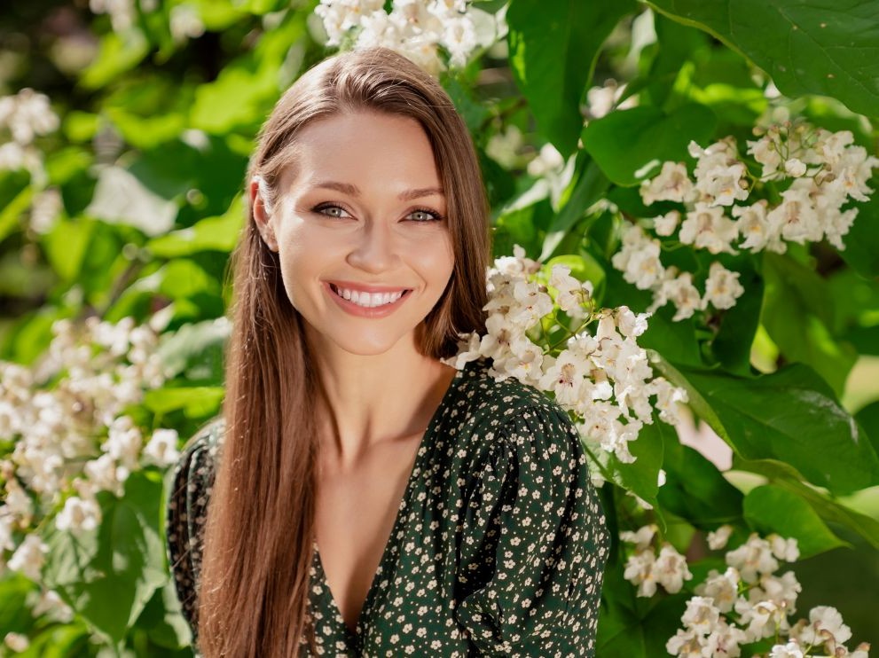Westside Behavioral Care About Us Westside Behavioral Care About Westside Section - Young woman with long brown hair smiling in front of lush green leaves and white blooming flowers on a sunny day.