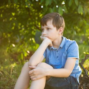 Westside Behavioral Care Anxiety Westside Behavioral Care Anxiety Section - Young boy in a blue shirt sitting on the ground in a shaded wooded area, resting his chin on his hand with a thoughtful expression.
