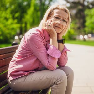 Westside Behavioral Care Depression Westside Behavioral Care Depression Section - Thoughtful older woman in glasses and a pink shirt sitting on a park bench, resting her face in her hands with a gentle expression.