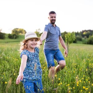 Westside Behavioral Care Parenting Skills Westside Behavioral Care Parenting Skills Section - Young girl wearing a sunhat and floral top running joyfully through a wildflower field while holding hands with her smiling father behind her.