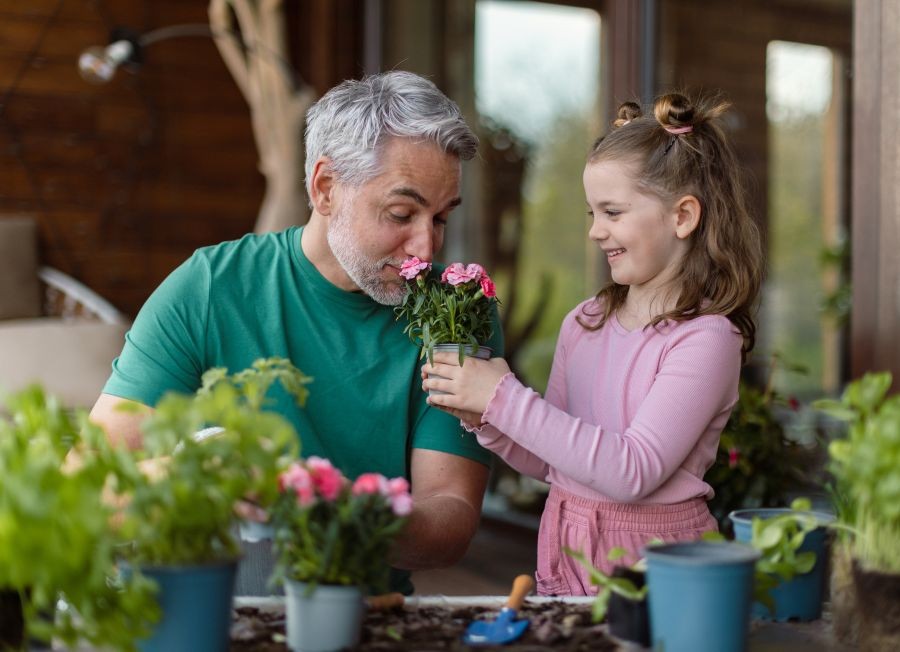 Westside Behavioral Care Patient Values Section - Smiling older man with gray hair smelling pink flowers held by a young girl, as they enjoy a gardening activity together on a sunny patio.