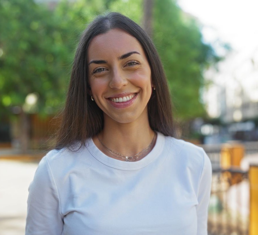 Westside Behavioral Care Therapist Values - Young woman with long dark hair smiling warmly at the camera, standing in a sunny urban park with trees and buildings in the background.