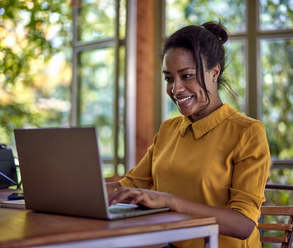 Westside Behavioral Care We Accept Insurance Westside Behavioral Care We Accept Insurance Section - Woman wearing a mustard yellow shirt smiling while working on a laptop at a wooden table in a bright room with large windows and green trees outside.